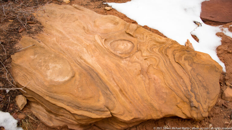 Orange sandstone shows circular bands and swirls beside a sharp edge of snow.