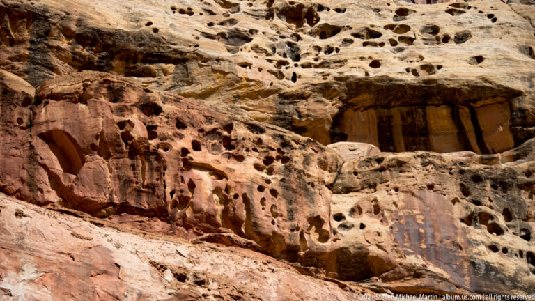 Rounded cavities and alcoves spread across pale and red sandstone layers in a broad rock face.