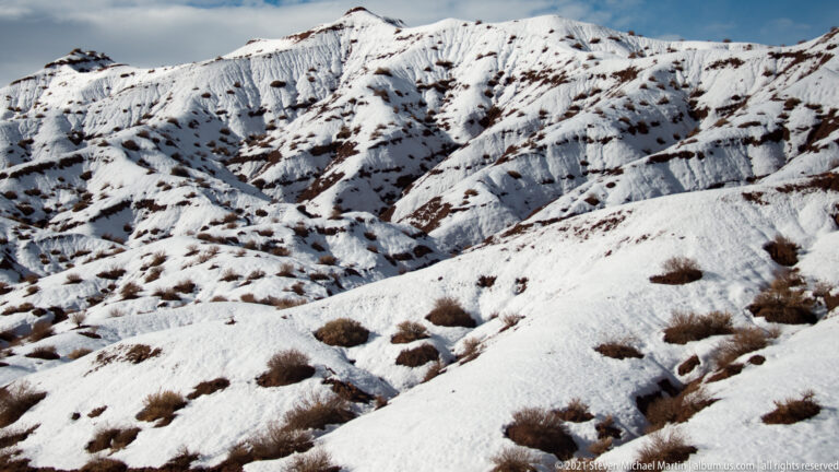 Snow-filled gullies descend a steep sandstone slope dotted with shrubs beneath a narrow strip of sky.