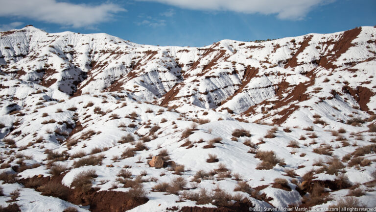 Snow covers a rolling hillside broken by dark-red sandstone ledges and scattered shrubs.