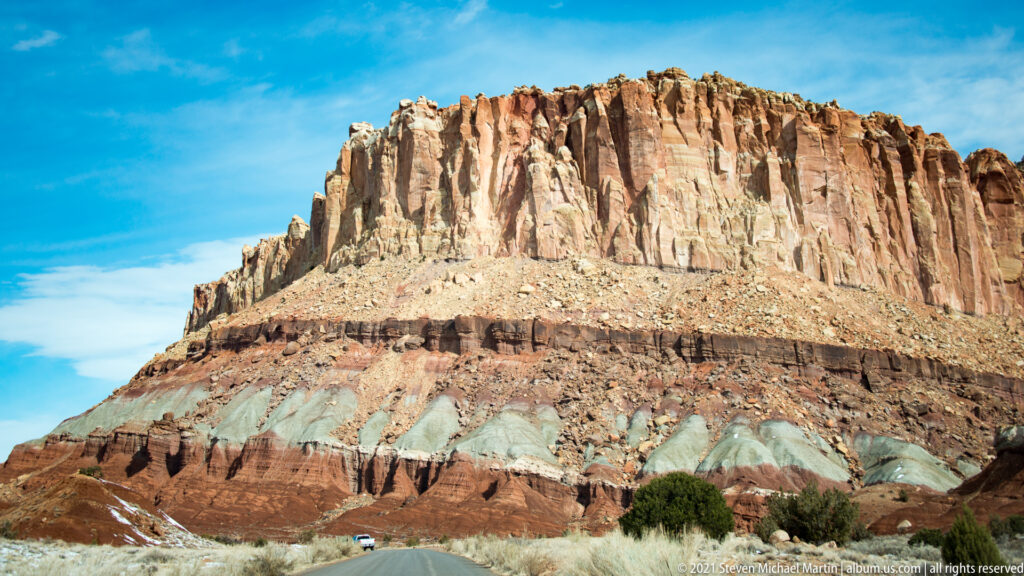 A high pale sandstone cliff rises above red and gray layers, a road and a small vehicle.