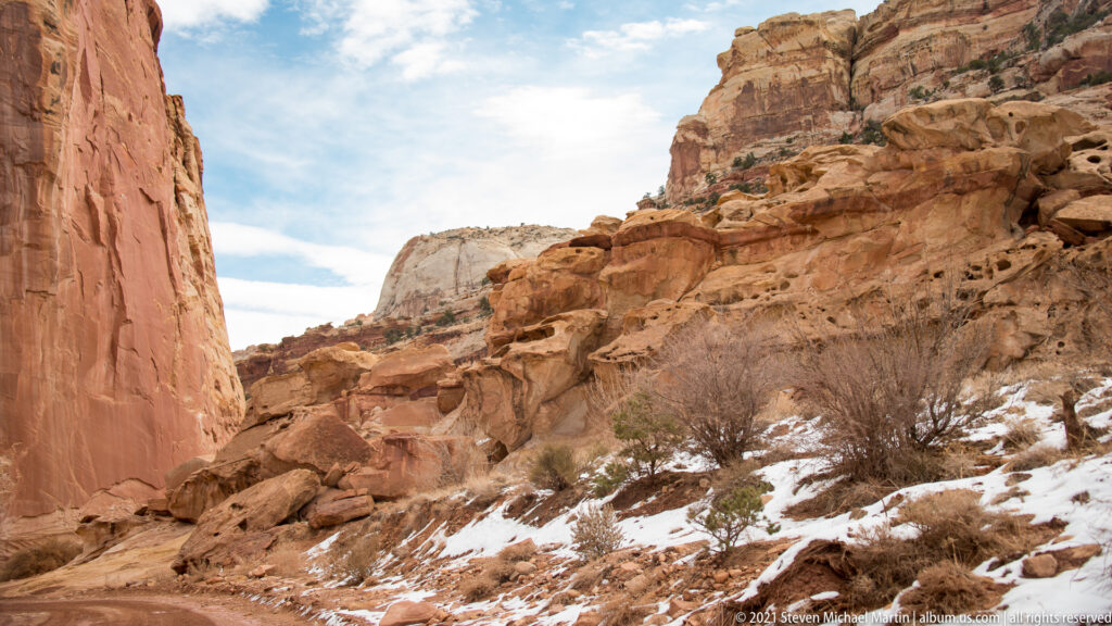 A dirt road curves between orange sandstone walls, with brush and patches of snow along its edges.