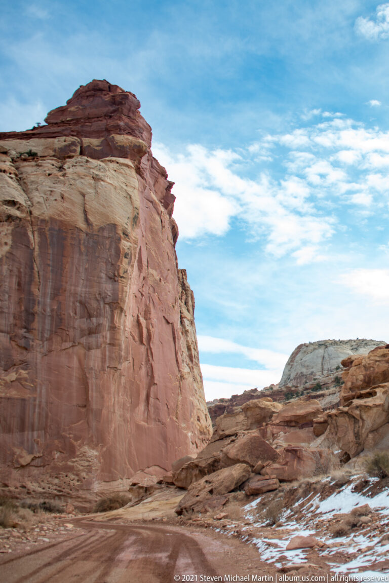 A towering red sandstone wall borders a low road cut, with a pale ridge and blue sky beyond.