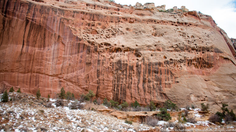 Dark runoff streaks mark a red sandstone wall beneath pale pitted rock, with snow at its base.