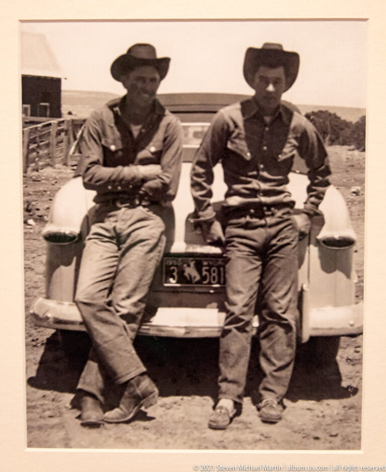 Two men wearing brimmed hats pose beside an older vehicle in a sepia photograph.
