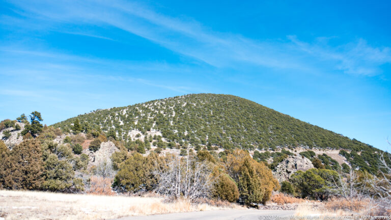 The snow-speckled, tree-covered cone rises beyond dry grass and a dense band of shrubs.