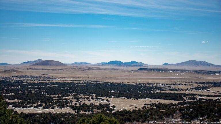 An elevated panorama looks across patchy woodland and open plains toward a distant line of volcanic hills.