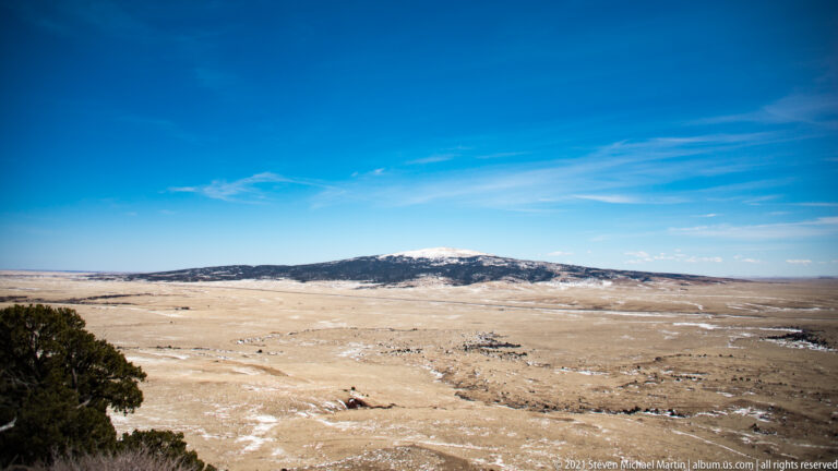 A pale winter plain reaches toward a snow-dusted ridge, with a dark evergreen tree at the left edge.