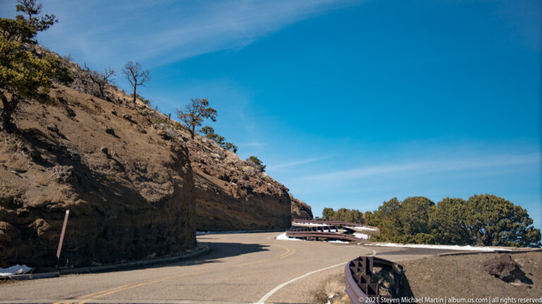 A paved road curves past a steep dark rock cut, with a guardrail and scrub trees along the shoulder.