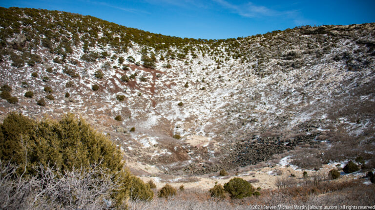 A steep crater slope is mottled with snow, dark rocks, and scattered low shrubs.