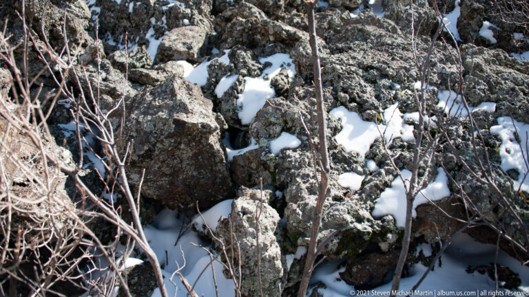 Angular dark boulders fill a rocky crevice where small patches of snow and bare stems collect.