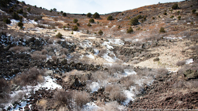 A shallow rocky drainage with patches of snow crosses a hillside dotted with low shrubs.