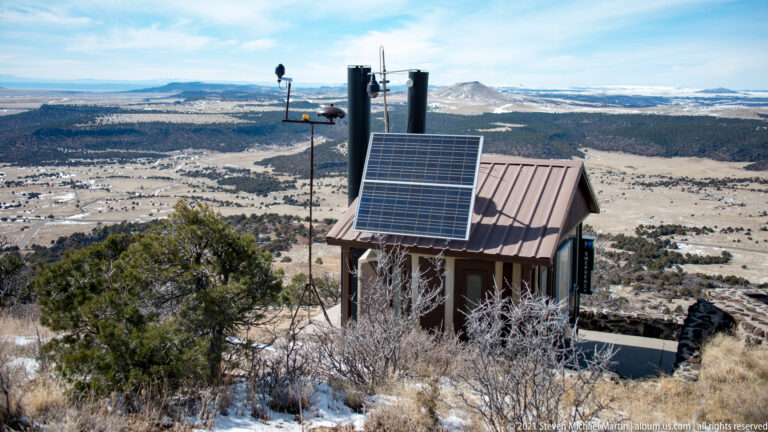 A small roofed structure with a solar panel and antennas stands on a wooded summit above a broad landscape.