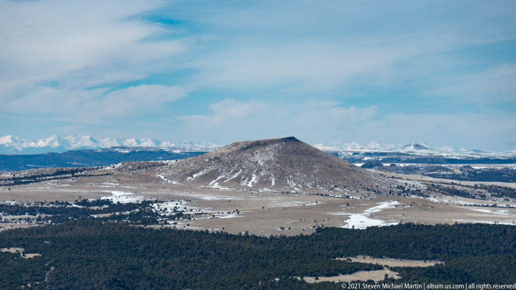 A rounded cinder cone rises beyond dark forest, with snow-covered mountains along the far horizon.