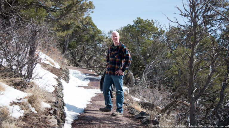 A visitor stands on a narrow trail edged with snow, rocks, and evergreen shrubs.