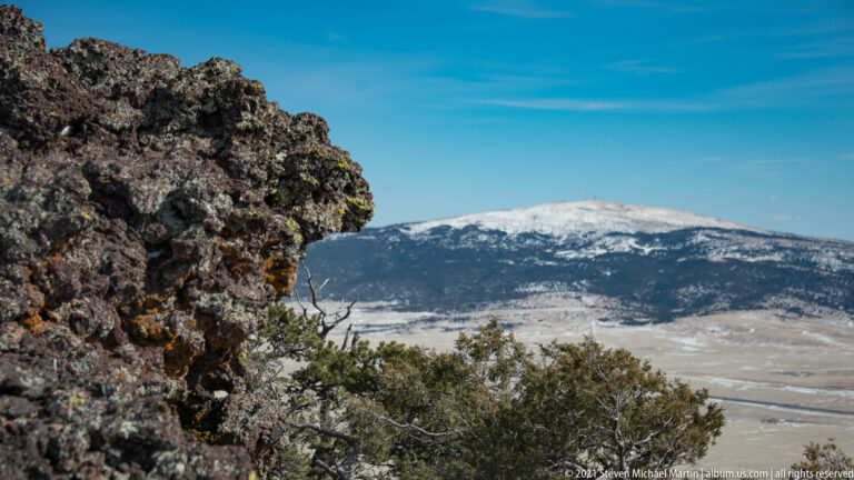A dark volcanic outcrop frames a snow-covered ridge beyond trees and an open valley.