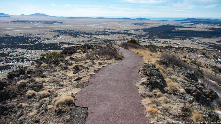 A paved trail descends a rocky volcanic slope toward a wide plain and distant mesas beneath a blue sky.