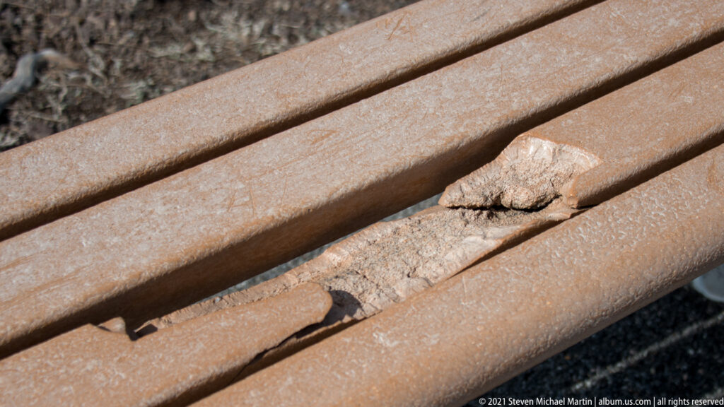 A rough pale rock fragment is lodged between parallel rust-colored metal bars, with vegetation blurred behind it.