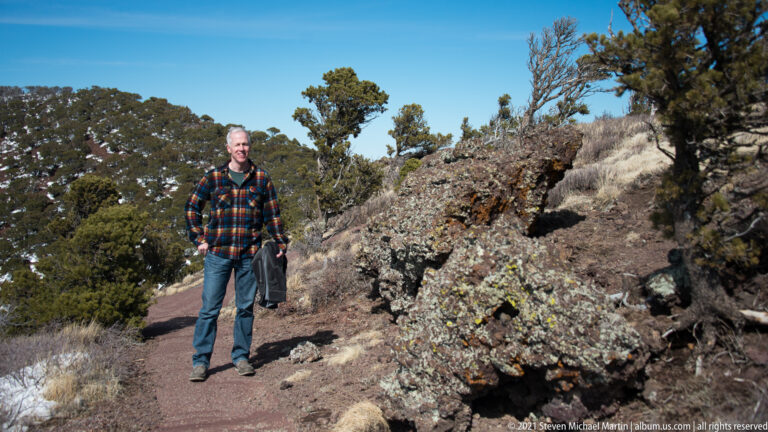 A man stands on a reddish trail beside a tall jagged rock outcrop surrounded by shrubs and grasses.