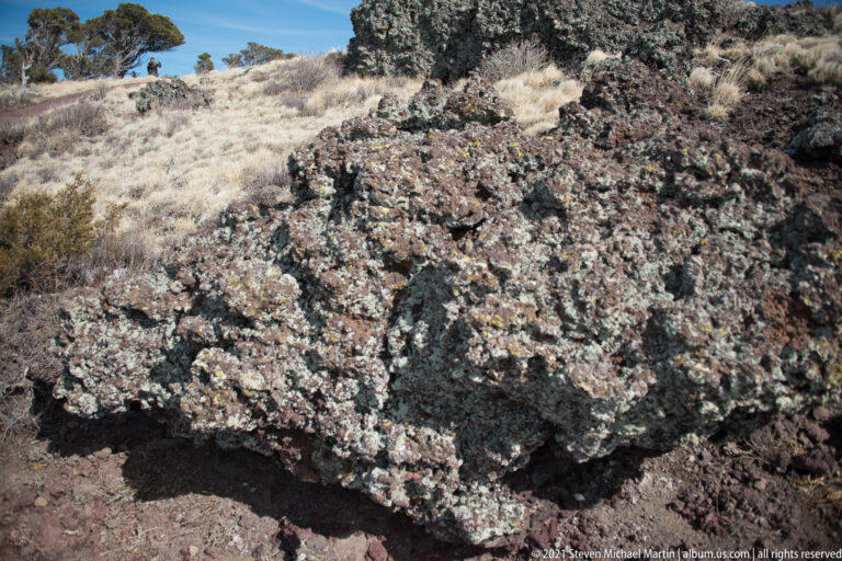 Fractured dark volcanic rocks fill the foreground, with pale grasses and shrubs growing in the gaps.