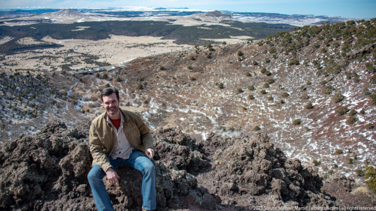 A seated man rests on dark summit rocks above a broad plain with patches of snow.