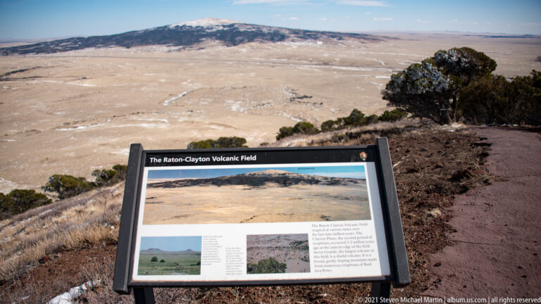 An interpretive panel labeled “The Raton-Clayton Volcanic Field” stands before a broad plain and distant hills.