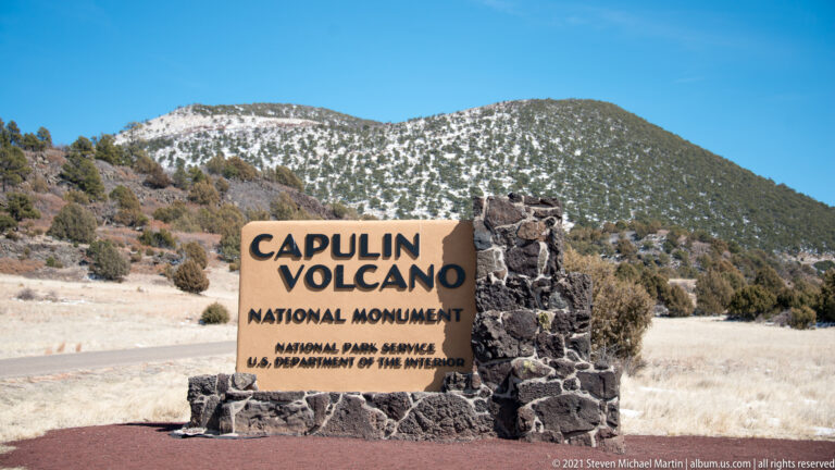 A stone monument sign reading Capulin Volcano National Monument stands before the snow-speckled volcanic cone.
