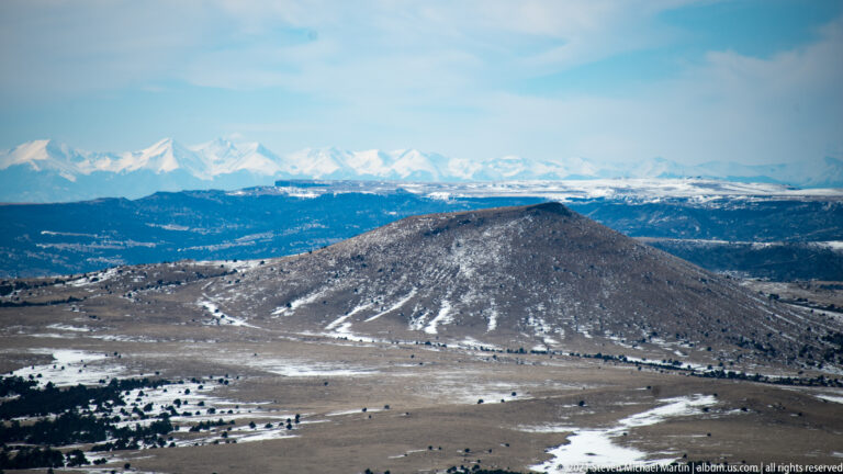 A dark, snow-streaked cinder cone rises from a broad plain, with snow-covered mountains on the horizon.
