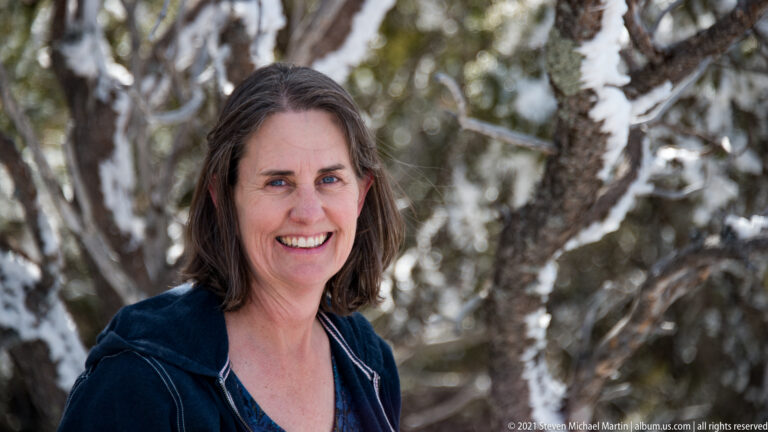 A smiling woman in a dark blue jacket poses before softly blurred winter trees.