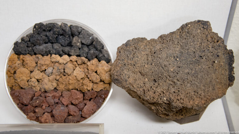 A large porous volcanic rock sits beside a round tray of smaller dark gray, brown, and reddish fragments.