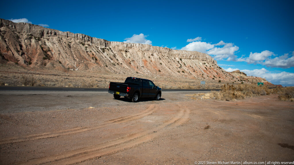 A dark pickup is parked beside a road below a tall, vertically eroded sandstone escarpment.