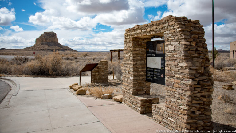 A stone gateway holds an information panel beside a paved path, with a freestanding sign and mesa beyond.
