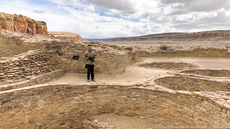 A visitor holding a camera stands among curved low walls and excavated spaces in a broad canyon site.