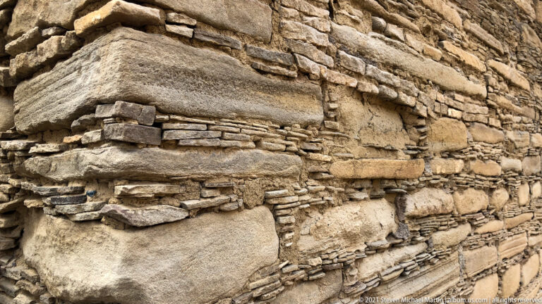 Broad pale slabs interrupt thin horizontal stone courses in a close view of a masonry wall.