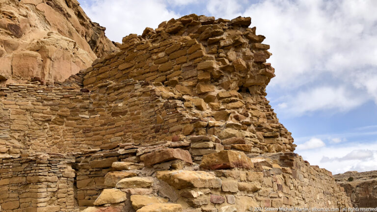 A tall masonry corner with broken upper stones rises above lower walls against a partly cloudy sky.