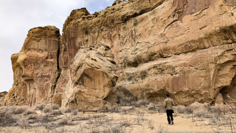 A person approaches a towering sandstone cliff with shrubs and a bench at its base.