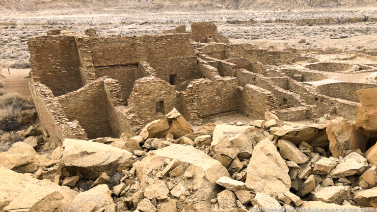 A large multiroom stone complex is seen from above, with rubble in front and open terrain beyond.