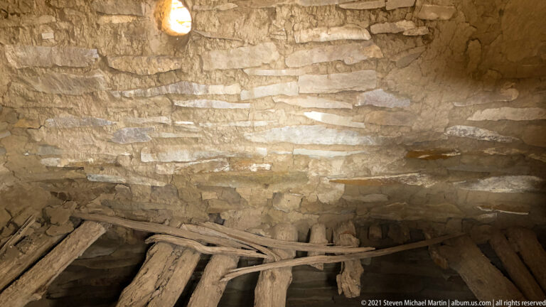 A stone chamber contains heavy timber beams beneath stepped masonry walls and a small high opening.