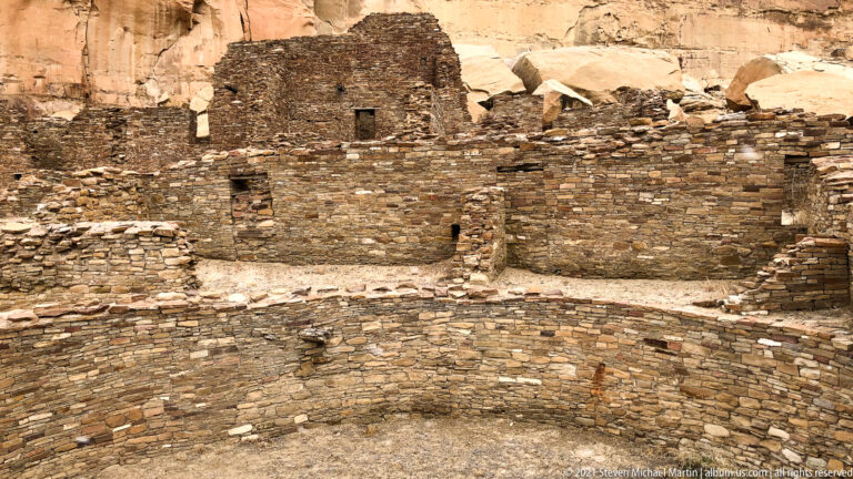 A curved stone enclosure sits before taller masonry rooms with small openings beneath a cliff.