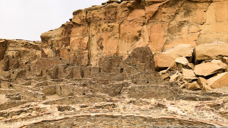 Low stone room blocks extend beneath a high sandstone cliff, with large fallen boulders at right.