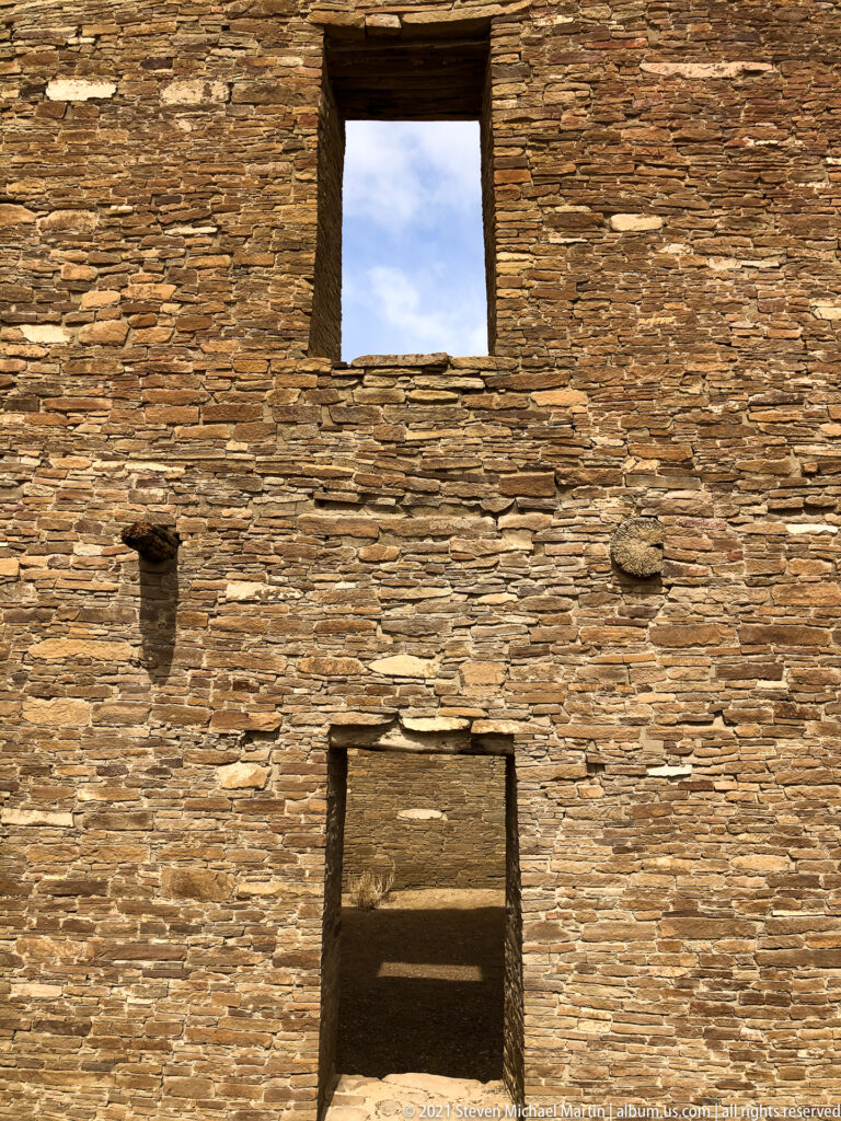 A straight-on stone façade has a large lower doorway, a smaller high opening, and a projecting beam.