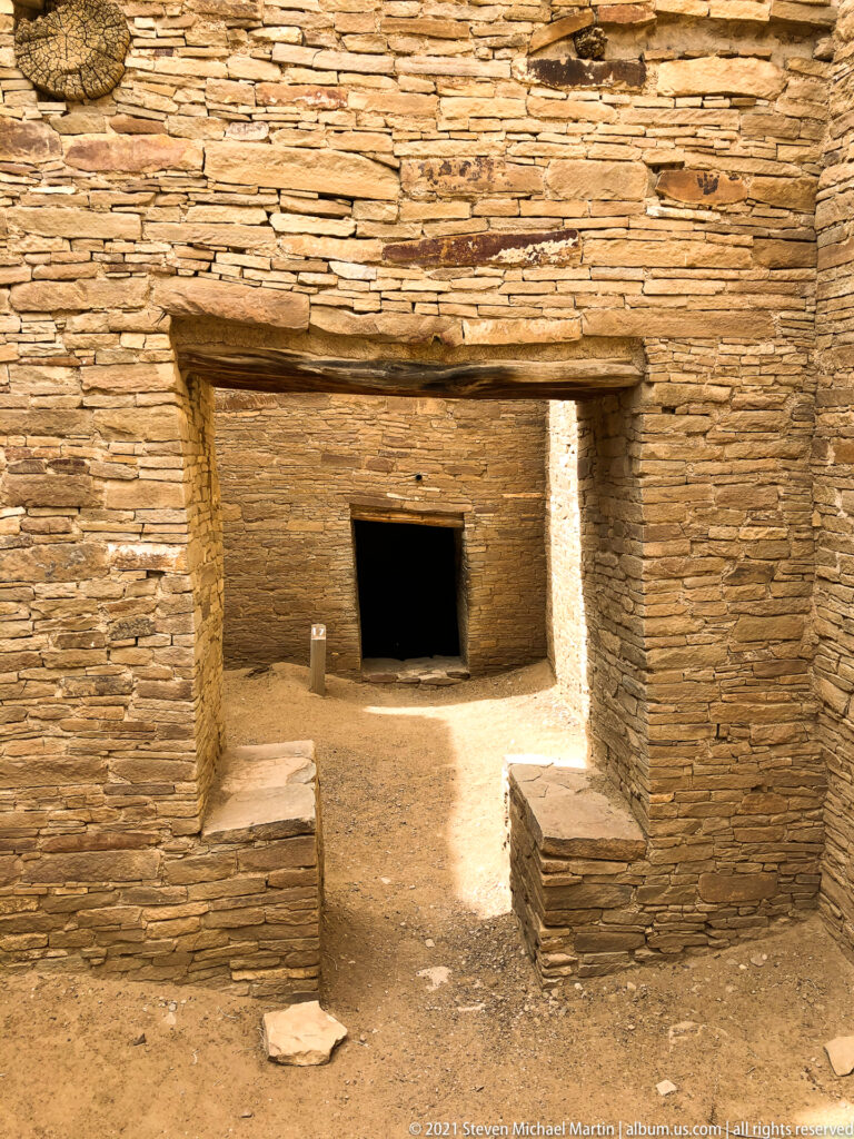 A broad lintel and stone jambs frame a sunlit entry aligned with a smaller dark doorway.