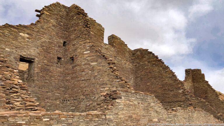 A long stone façade with small openings and a broken stepped profile rises beneath cloudy sky.