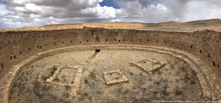 An elevated view looks into a circular stone kiva with small wall openings and low structures on its floor.