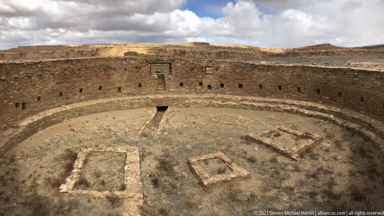 A curved kiva wall with small openings surrounds a dry floor and several low stone features.