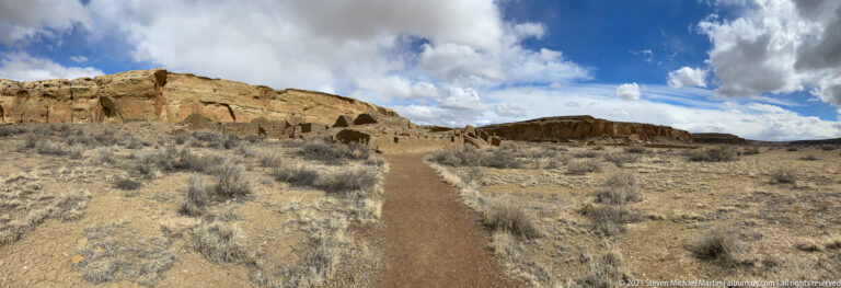 A dirt path leads through dry brush toward low stone structures beneath layered sandstone cliffs and clouds.