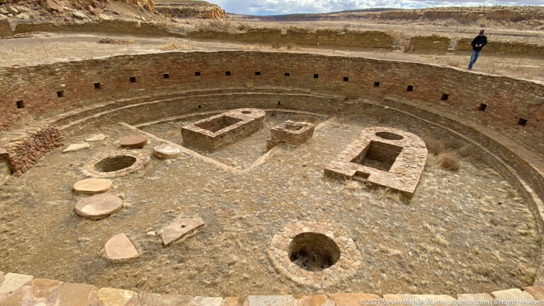A visitor stands on the rim of a circular kiva above low stone features inside its curved walls.