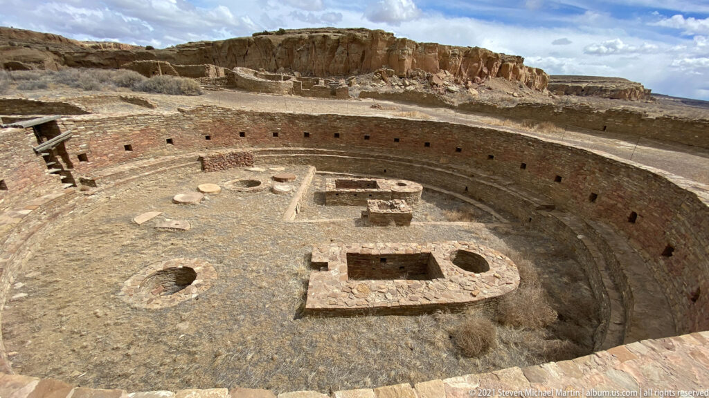 A broad circular kiva fills the foreground below a sandstone cliff, with low masonry features inside.