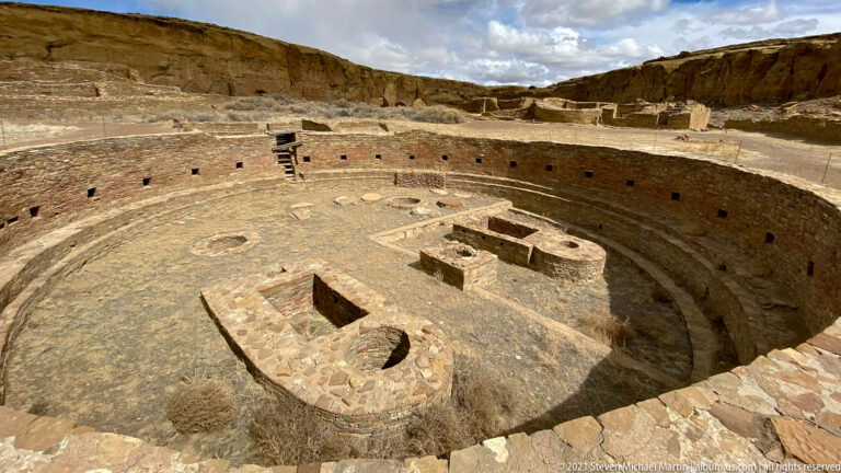 A low stone structure with rounded ends sits inside a circular kiva, with more ruins and cliffs beyond.