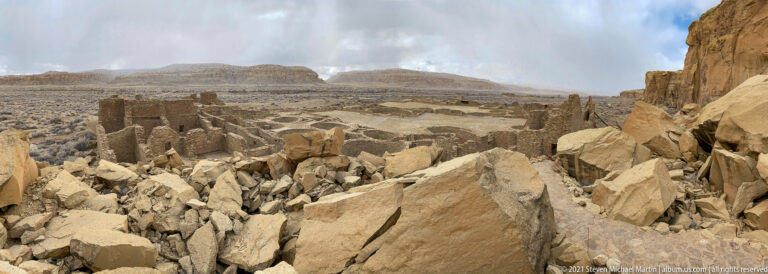 Large broken sandstone blocks lie before standing room walls and foundations across an open canyon.